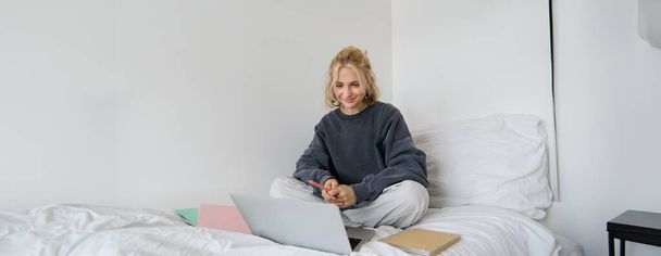 Porträt einer glücklichen blonden Frau, die von zu Hause aus arbeitet und mit Laptop und Notizbuch im Bett sitzt. Student macht Hausaufgaben im Schlafzimmer, verbindet sich per Videochat mit dem Online-Unterricht. - Foto, Bild