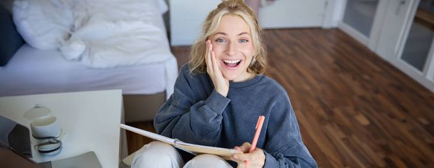 Porträt einer glücklichen, jungen blonden Frau, die mit Notizbuch und Stift im Schlafzimmer sitzt, lacht und lächelt, im Tagebuch schreibt, Hausaufgaben macht und lächelt. - Foto, Bild