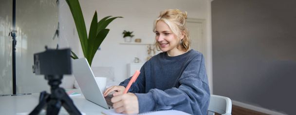 Портрет молодої блондинки усміхненої жінки, яка навчається вдома, концепція дистанційної освіти, підключається до онлайн-курсу або уроку, пишучи в блокноті. - Фото, зображення