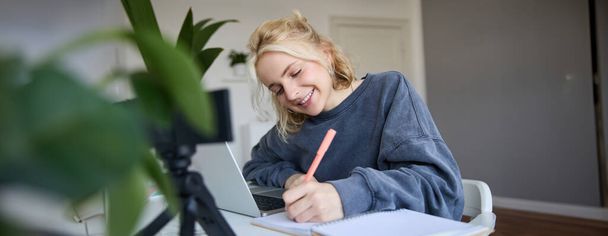 Πορτρέτο της νεαρής γυναίκας, lifestyle blogger, μαγνητοσκόπηση του εαυτού της, σημειώσεις, γράψιμο σε ημερολόγιο, να κάθεται μπροστά από το laptop σε ένα δωμάτιο και να σπουδάζει. - Φωτογραφία, εικόνα