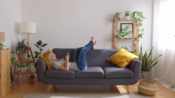 Täysi kulma näkymä onnellinen nuori valkoihoinen nainen käyttää matkapuhelinta, kun makaa sohvalla kotona. - Materiaali, video