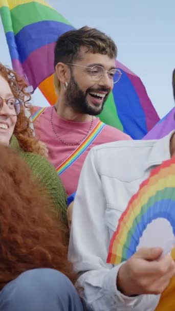 Vertical. Joyful diverse people gen z gathered group sitting celebrating LGBT party together. Laughing friends community enjoying gay pride day having fun outdoor. Flags and rainbow fans accessories - Footage, Video