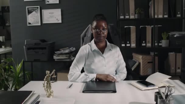 Ritratto medio di una seria avvocato afroamericana in occhiali e abbigliamento da ufficio che posa a tavola in un ufficio moderno e guarda la macchina fotografica - Filmati, video