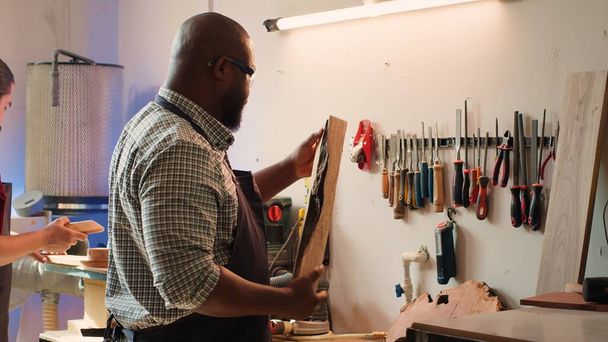 Homem afro-americano na loja de marcenaria inspecionando madeira antes de montar móveis, verificando se há arranhões. Fabricante de armários avalia bloco de madeira, garantindo que atende aos padrões de qualidade, câmera A - Foto, Imagem