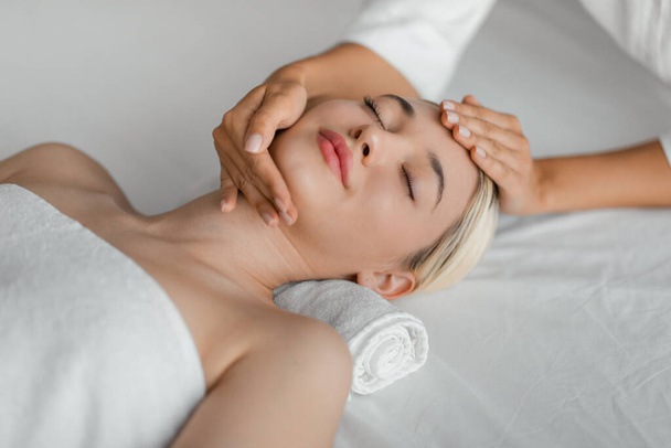 Een vrouw ligt op een kuuroord terwijl een professional zachtjes haar gezicht masseert, gericht op drukpunten en gebieden van spanning, bovenaanzicht - Foto, afbeelding