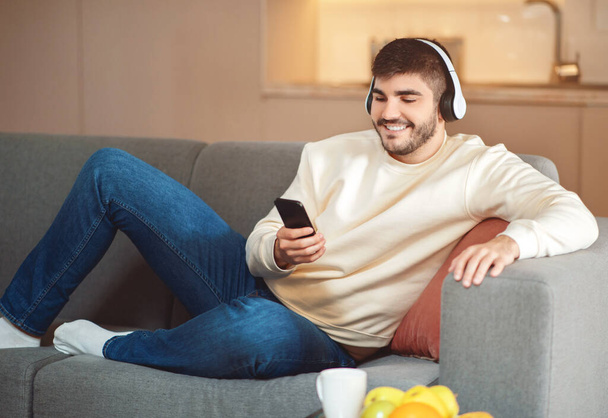 Um jovem sorridente descansa confortavelmente em um sofá cinza em um ambiente acolhedor sala de estar. Ele está usando roupas casuais e fones de ouvido, e está segurando um smartphone, possivelmente escolhendo música. - Foto, Imagem