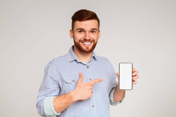 Sorrindo milenar homem segurando mais recente celular magro com tela branca e apontando para ele, panorama, espaço livre
 - Foto, Imagem
