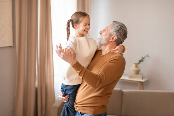 Un hombre mayor está de pie, sosteniendo a una niña sonriente en sus brazos. Ambos parecen felices y conectados, mostrando un momento conmovedor de amor familiar. - Foto, Imagen