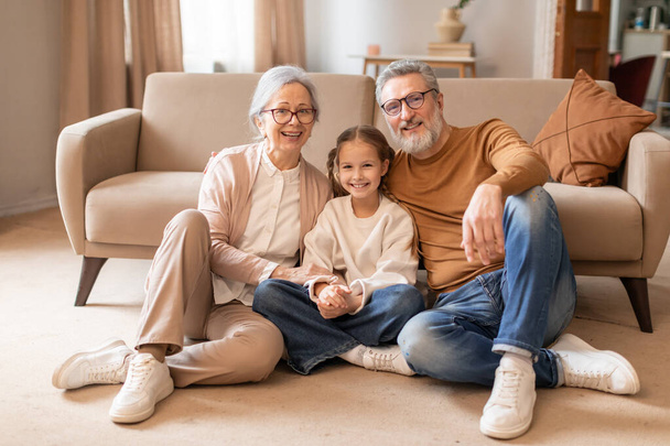 Ein fröhliches älteres Ehepaar und seine junge Enkelin sitzen eng beieinander auf dem Boden eines gemütlichen Wohnzimmers und strahlen Wärme und familiäre Zweisamkeit aus.. - Foto, Bild
