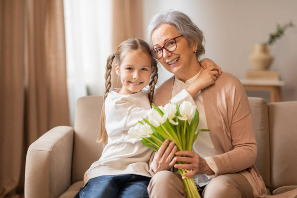 Een vrolijke oma met bril en haar kleindochter, met staartjes, poseren samen op een beige bank. De kleindochter houdt een boeket verse witte tulpen vast. - Foto, afbeelding