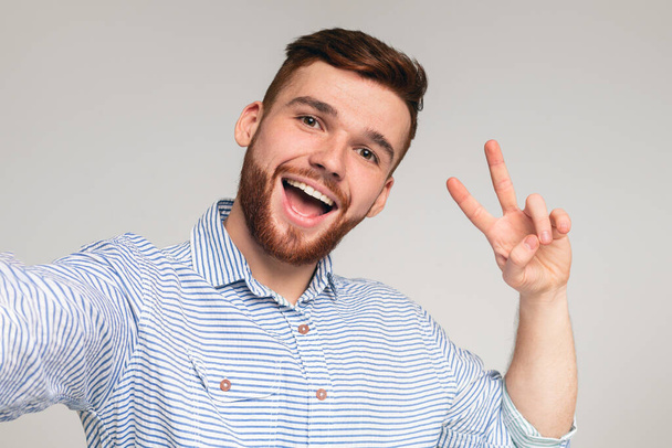 Young millenial man showing V - sign and making selfie on beige background, panorama - Photo, Image
