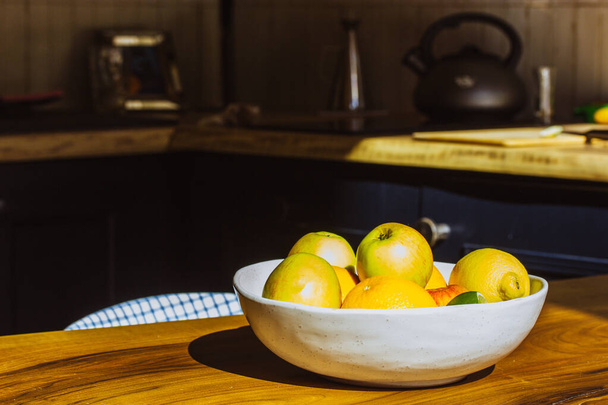 Ένα πιάτο με φρέσκα φρούτα εποχής στέκεται σε ένα ξύλινο τραπέζι σε μια ρουστίκ άνετη κομψή κουζίνα. Υγιεινή διατροφή, διατροφή, βιταμίνες για έναν υγιεινό τρόπο ζωής. Μήλα, πορτοκάλια, λεμόνια, εσπεριδοειδή σε πιατέλα.  - Φωτογραφία, εικόνα