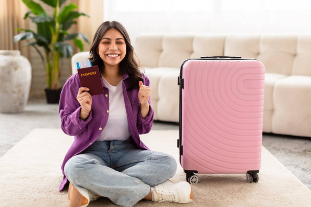 Улыбающаяся женщина с Ближнего Востока сидит на полу со скрещенными ногами, держа паспорт, с упакованным чемоданом, готовым к путешествию - Фото, изображение