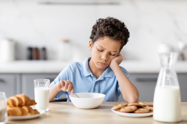 Sur cette image, un garçon afro-américain s'ennuie assis à la table de la cuisine, désintéressé par son petit déjeuner, mettant en évidence un scénario commun du matin de la famille - Photo, image