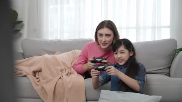 Happy girl playing game and sitting while mother lie on sofa. Caucasian parent and attractive smart girl spending time together while holding joystick and focus on winning games. Lifestyle. Pedagogy. - Footage, Video