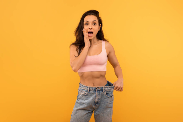 A young woman stands with a shocked expression on her face, her hand touching her cheek, wearing a casual pink crop top paired with oversized blue denim jeans - Photo, Image