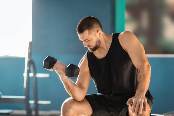 Un homme est assis sur un banc de gym, tenant une paire d'haltères dans ses mains. Il semble concentré et concentré sur sa routine d'entraînement, montrant sa détermination et son engagement envers la condition physique.. - Photo, image
