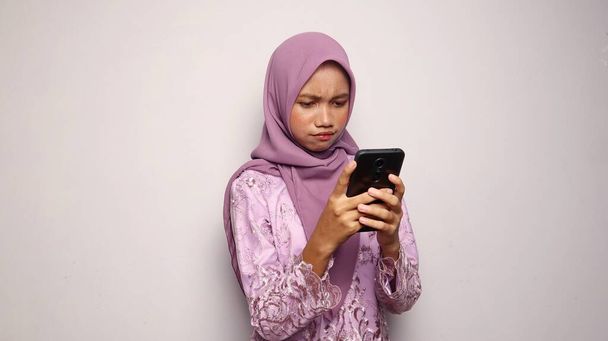 Krásné indonéské dospívající dívka na sobě kebaya a hidžáb držení a při pohledu na smartphone na izolovaném bílém pozadí - Fotografie, Obrázek