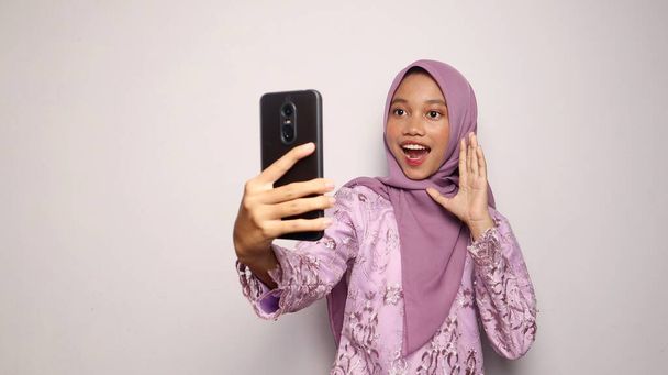 インドネシアの十代の少女は,ケバヤとヒジャブを着用して,孤立した白い背景にスマートフォンセルフィーを保持しています - 写真・画像
