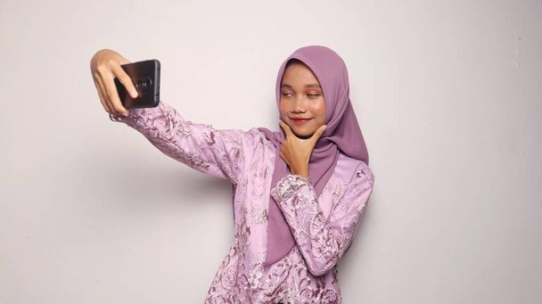 インドネシアの十代の少女は,ケバヤとヒジャブを着用して,孤立した白い背景にスマートフォンセルフィーを保持しています - 写真・画像