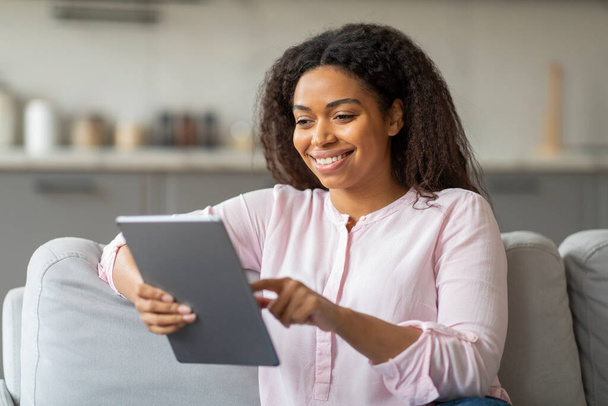 Přístupná černá žena drží tablet, její úsměv naznačuje požitek nebo uspokojení z obsahu ona prohlíží doma - Fotografie, Obrázek