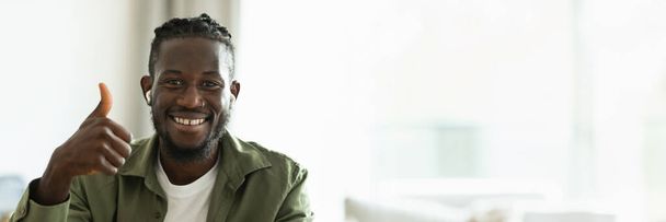 Porträt eines positiven afrikanisch-amerikanischen Freiberuflers, der aus der Ferne arbeitet und mit erhobenem Daumen gestikuliert, während er zu Hause im Büro sitzt. Ich mag Freiberuflichkeit und Unternehmertum, Panorama mit Kopierraum - Foto, Bild