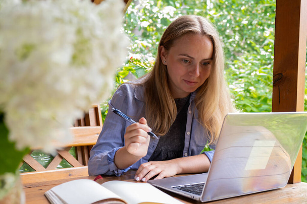 Uczennica ma wykształcenie online w ogrodzie drewniana alkowa. Blondynka siedząca na zewnątrz, pracująca na laptopie, dzwoniąca wideo. Jedność z naturą - Zdjęcie, obraz
