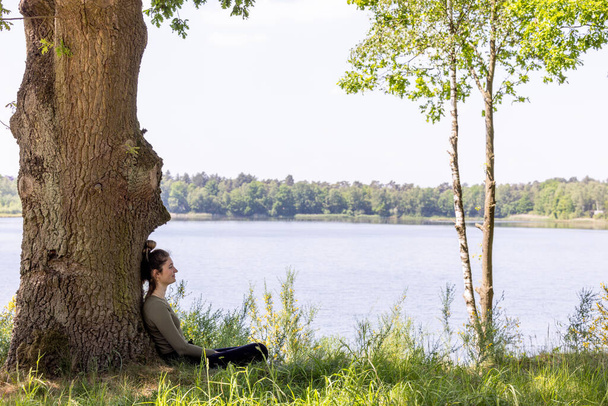 Egy csendes nyugalom képe, amint egy nő ül egy fa erős törzse alatt. A tó a háttérben, keretezett zöld lombozat, kiemeli az érzés, a magány és a kapcsolat - Fotó, kép