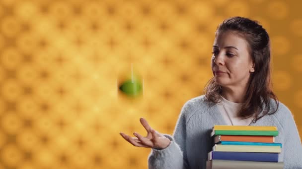 Szczęśliwa kobieta trzymająca książki i bawiąca się jabłkiem, przekazująca radość z czytania i jedzenia zdrowego, studyjnego tła. Wesoła osoba z powieściami i świeżymi owocami, koncepcja dobrego stylu życia, kamera A - Materiał filmowy, wideo
