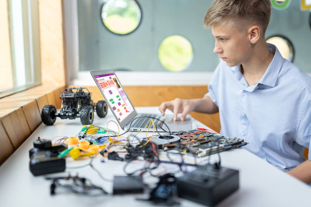 Schoolboy watching code on screen and using touch pad of laptop while learn technology in STEM class. On table put laptop, controller, electric wire, battery charger, and robotic vehicle. Edification. - Photo, Image