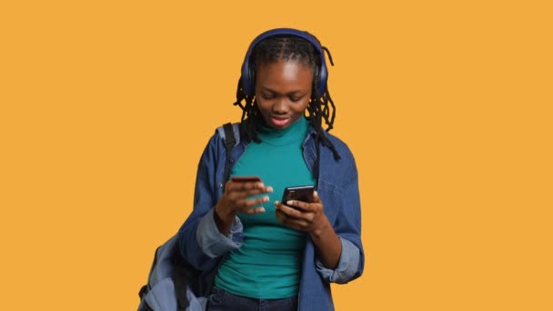 Afroamerikanischer Teenager, der eine Zahlungsmethode auf einer Webseite hinzufügt, isoliert über den Studiohintergrund. Junges Mädchen trägt Kopfhörer, erledigt Internet-Einkäufe, tippt Kreditkarteninformationen auf das Telefon, Kamera A - Filmmaterial, Video