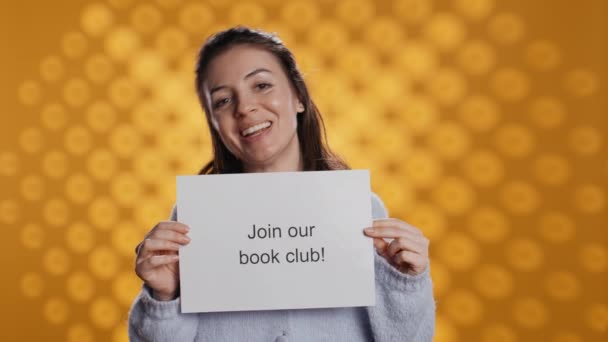 Cheerful book club president holds message urging people to join them, talking about importance of lecture, studio background. Smiling woman inviting bookworms to enlist in her association, camera B - Footage, Video