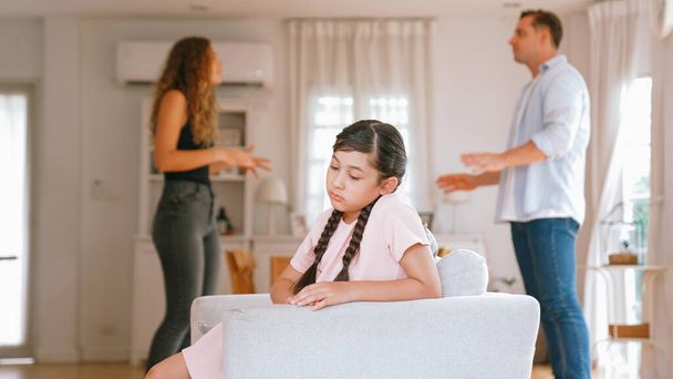 Annoyed and unhappy young girl sitting on sofa trapped in middle of tension by her parent argument in living room. Unhealthy domestic lifestyle and traumatic childhood develop to depression Synchronos - Photo, Image