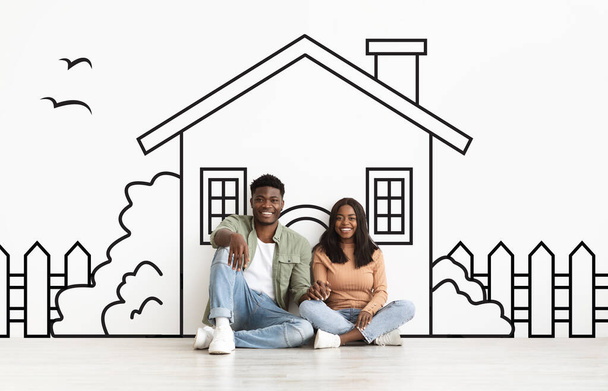 Afroamerikanische Ehepartner sitzen auf dem Boden, einander gegenüber, vor ihnen die Zeichnung eines Hauses. Sie scheinen die Skizze zu diskutieren oder zu bewundern. - Foto, Bild