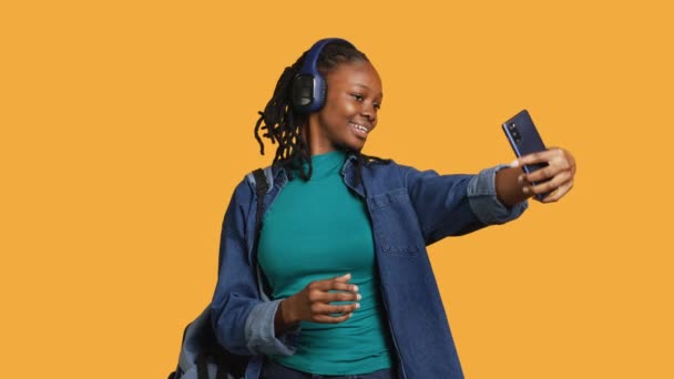 Τζόλι έφηβος που χρησιμοποιεί κινητό για να βγάλει σέλφι κάνοντας σήμα νίκης. Χαμογελώντας κορίτσι BIPOC λήψη φωτογραφιών χρησιμοποιώντας το κινητό τηλέφωνο, κάνει gesturing νίκη, ακούγοντας μουσική, στούντιο φόντο, κάμερα A - Πλάνα, βίντεο