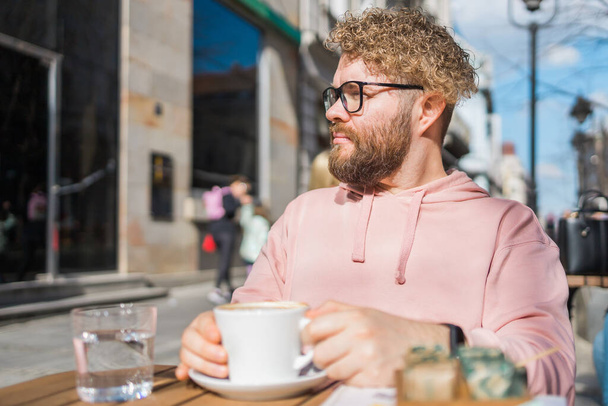 Milenium brodaty człowiek jedzący śniadanie przy stole ulicznej kawiarni w wiosenny dzień, pijący ciepłe cappuccino. Wiosenny taras restauracji. - Zdjęcie, obraz