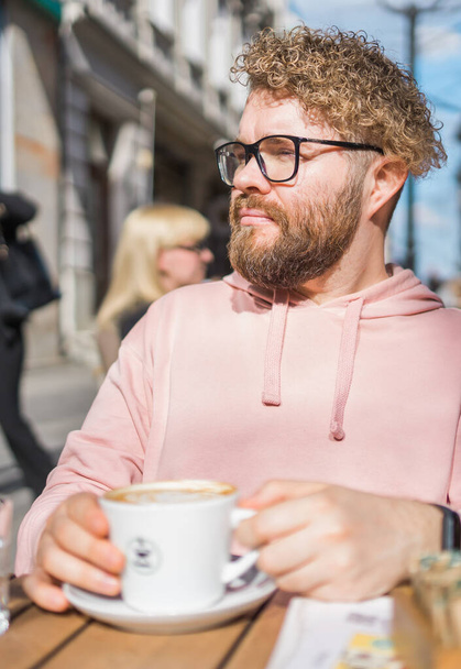 Millennial bearded man having breakfast at table of street cafe on spring day, drinking warm cappuccino. Spring restaurant terrace. - Photo, Image
