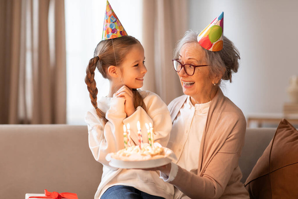 Una nonna allegra che indossa un cappello da festa presenta una torta di compleanno con candele accese a una giovane ragazza, anche in un cappello da festa, in quanto condividono uno sguardo amorevole e un caldo - Foto, immagini
