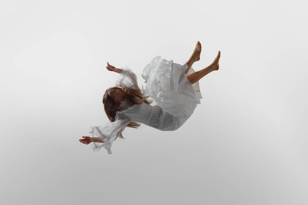 Femme en robe blanche fluide suspendue dans l'air dans une pose élégante et gracieuse sur fond de studio blanc. Une photo dynamique. Concept de beauté, élégance féminine et pureté, rêve et réalité. - Photo, image