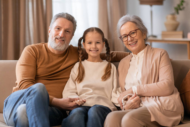 Une scène chaleureuse capture une jeune fille assise entre ses grands-parents, partageant un moment de joie à l'intérieur de la maison - Photo, image