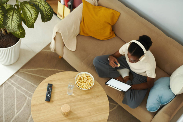 Jovem negra sentada no sofá, comendo pipocas e assistindo show no tablet - Foto, Imagem