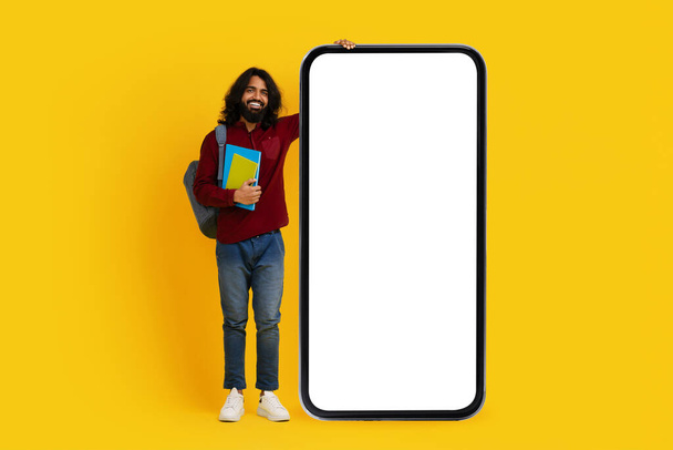 Homme indien joyeux avec un étudiant de barbe se tient sur le côté gauche, tenant des livres colorés et s'appuyant sur un massif, smartphone écran vide - Photo, image