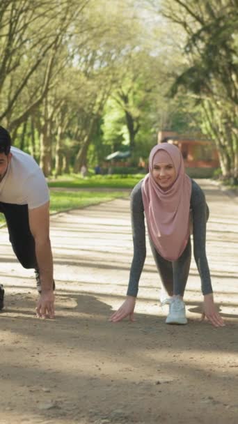 Vertikální obrazovka: Mladý muslimský pár ve sportovním oblečení stojí ve výchozí poloze pro běh v zeleném letním parku. Muž a žena v hidžábu se připravují na ranní cvičení venku - Záběry, video