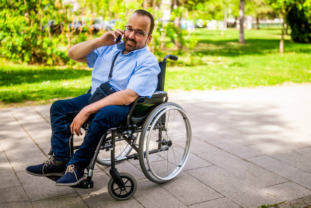 Portret van een gelukkige man in een rolstoel. Hij geniet van de zonnige dag in het stadspark en telefoneert. - Foto, afbeelding