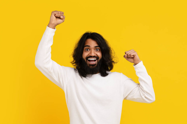 Parrakas nuori intialainen mies, jolla on pitkät hiukset, huokuu jännitystä ja iloa, kun hän nostaa nyrkkinsä voittoisaan eleeseen, keltainen tausta välittää voiton ja riemun tunteen.. - Valokuva, kuva
