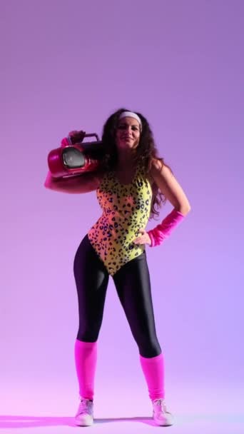 Fröhliche und freundliche kaukasische Frau aus den 80er Jahren, die Stereoanlage in der Hand hält, tanzt und Spaß zu Musik auf violettem Hintergrund hat. Vertikales Video. - Filmmaterial, Video