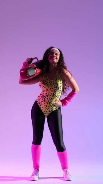 Wesoła biała kobieta w retro sportowym stroju z lat 90-tych, tańcząca do muzyki z boomboxem na ramieniu na fioletowym tle. Pionowe wideo. - Materiał filmowy, wideo