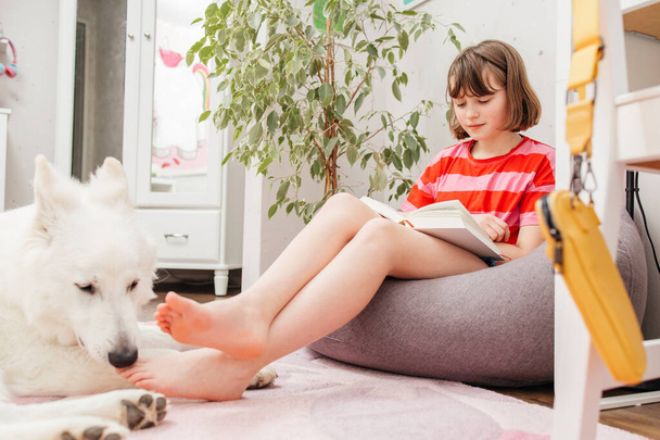 Une adolescente s'assoit sur un pouf dans sa chambre et lit un livre, à côté d'elle se trouve un chien, un berger suisse blanc. Lire des livres comme passe-temps. - Photo, image
