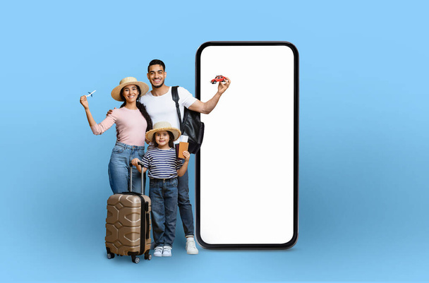 Una familia con equipo de viaje y un coche de juguete junto a un teléfono inteligente maqueta, que ilustra la tecnología de planificación de viajes, aislado sobre un fondo azul - Foto, imagen