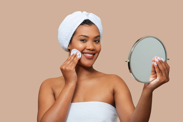 A young, cheerful woman with a towel wrapped around her hair is gently applying facial toner to her cheek. She holds a small, round mirror in one hand to ensure precise application - Photo, Image